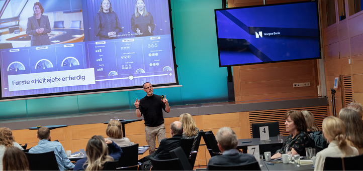 A man standing and talking in front of a sitting audience with a large screen in the background