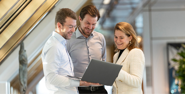 Two male and one female employee standing and looking at an open laptop that one of the men is holding in his hand