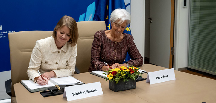 Two women sitting at a desk with a flower arrangement, each signing a contract