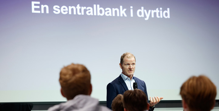 A man stands and talks in front of young people with the text "A central bank in a time of crisis"(in Norwegian) on a screen in the background